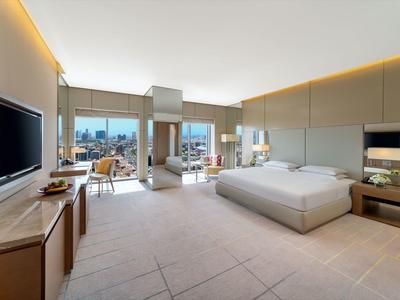 Ruime hotelkamer met tweepersoonsbed, zithoek en grote ramen met uitzicht op de stad.