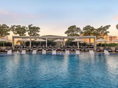 Großer Hotelpool mit Liegestühlen und Sonnenschutz, umgeben von Bäumen und Gebäuden.