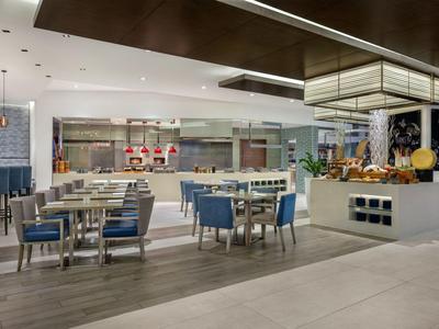 Moderní hotelová restaurace s modrými židlemi, bílými stoly a snídaňovým bufetem.