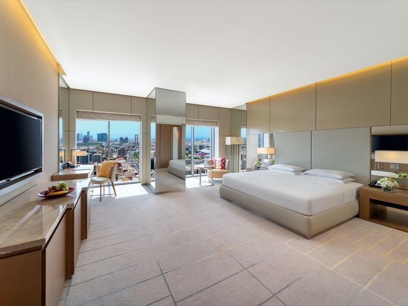 Ruime hotelkamer met tweepersoonsbed, zithoek en grote ramen met uitzicht op de stad.