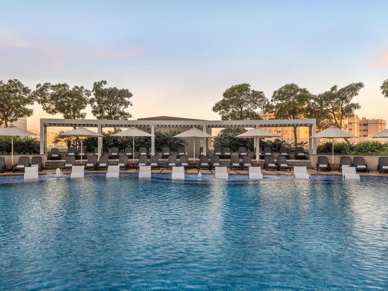 Groot hotelzwembad met ligstoelen en zonneschermen omringd door bomen en gebouwen.