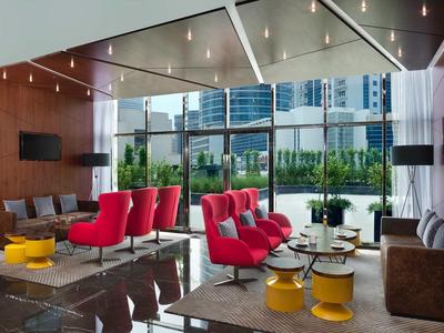 Moderna sala de hotel con sillones rojos, grandes ventanas y vista a edificios de la ciudad al fondo.