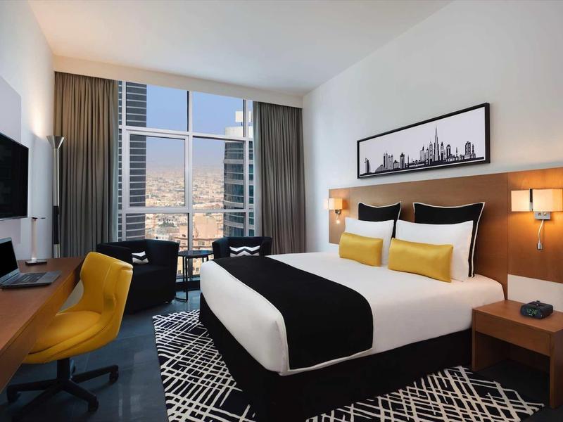 Habitación de hotel moderna con cama cómoda, escritorio, televisor y vista a la ciudad por ventana grande.