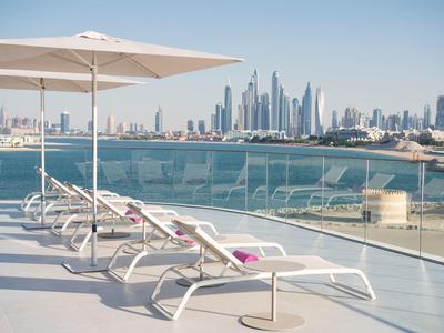 Weiße Liegestühle unter Sonnenschirm auf Terrasse mit Blick auf Meer und moderne Skyline.
