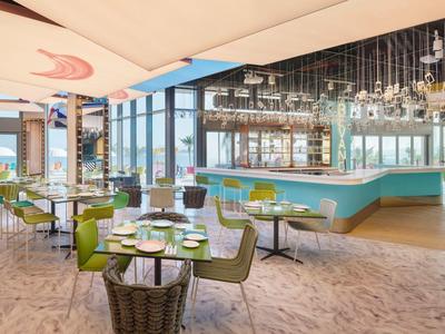 Modernes Restaurant mit großen Fenstern, grünen Stühlen, hellem Boden und eleganter Bar.