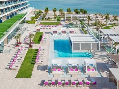 Moderner Hotelpool mit weißen Liegen, rosa Polstern, Palmen und Blick aufs blaue Meer.