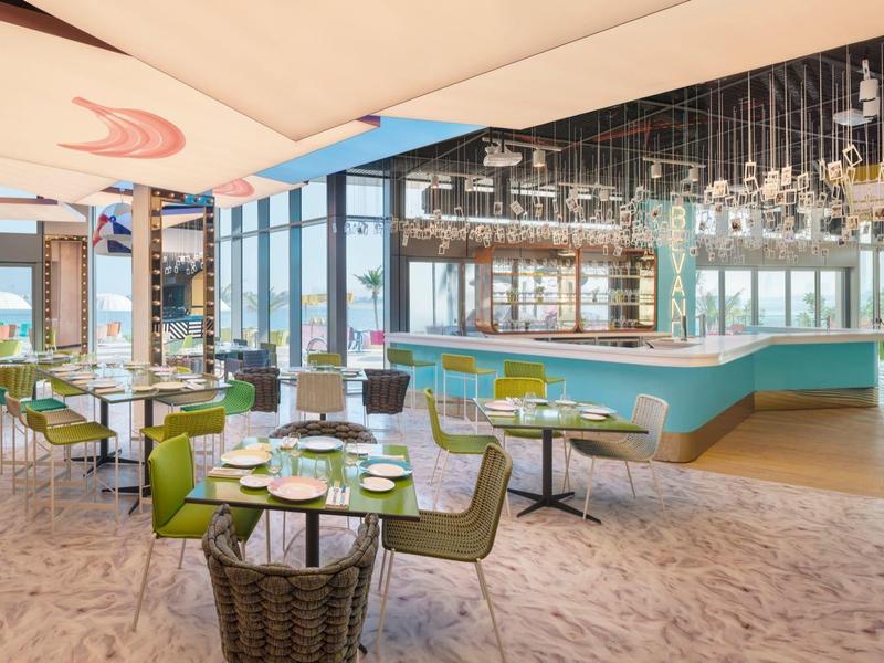 Modernes Restaurant mit großen Fenstern, grünen Stühlen, hellem Boden und eleganter Bar.