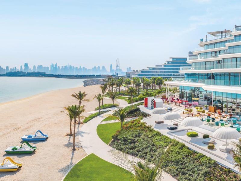 Strand mit Liegestühlen, Palmen und modernem Hotel am Wasser bei klarem Himmel.