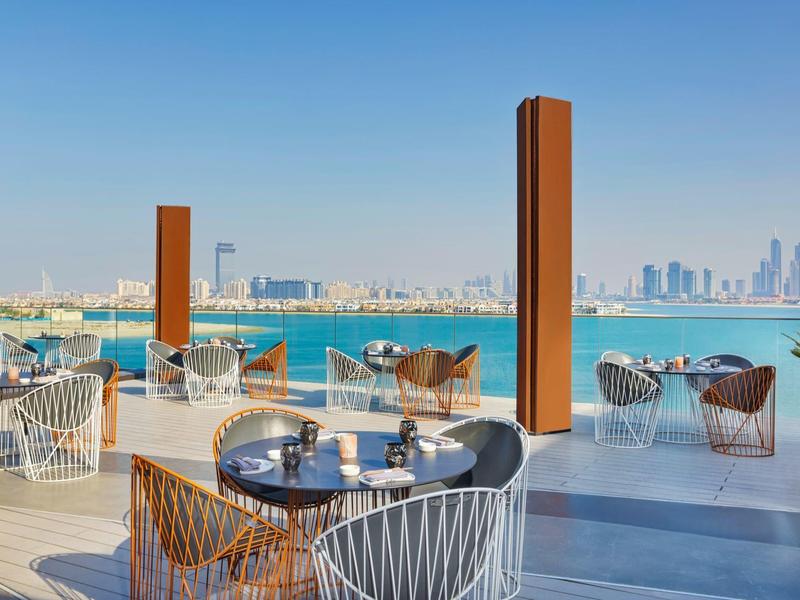 Terrasse mit modernen Stühlen und Tischen, Blick auf blaues Meer und entfernte Stadt mit Wolkenkratzern.