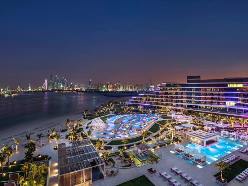 Luxushotel mit beleuchteten Pools und Stadtlichtern am Wasser bei Nacht.