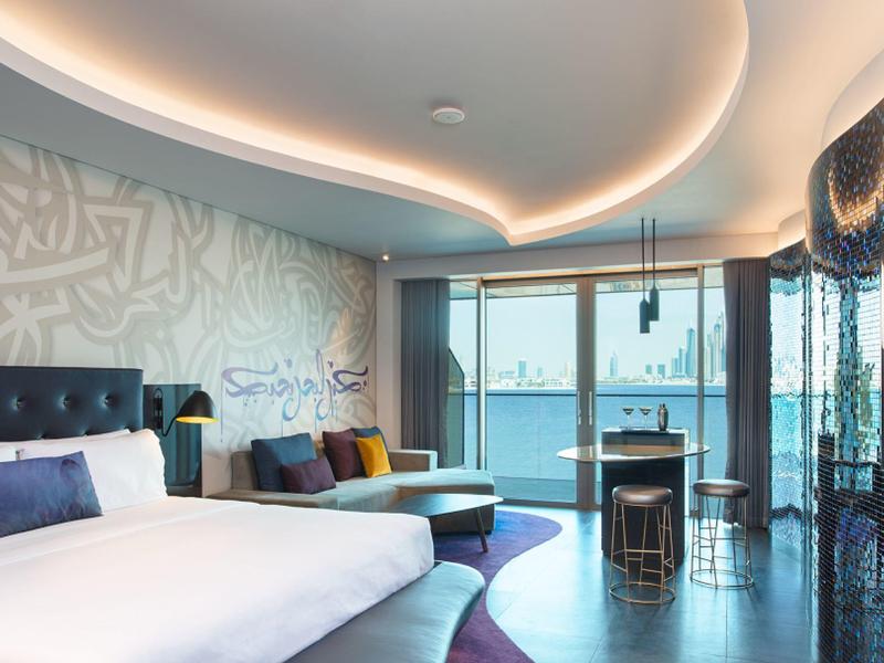 Modernes Hotelzimmer mit großem Bett, bunter Couch, Glasfensterfront und Meeresblick.