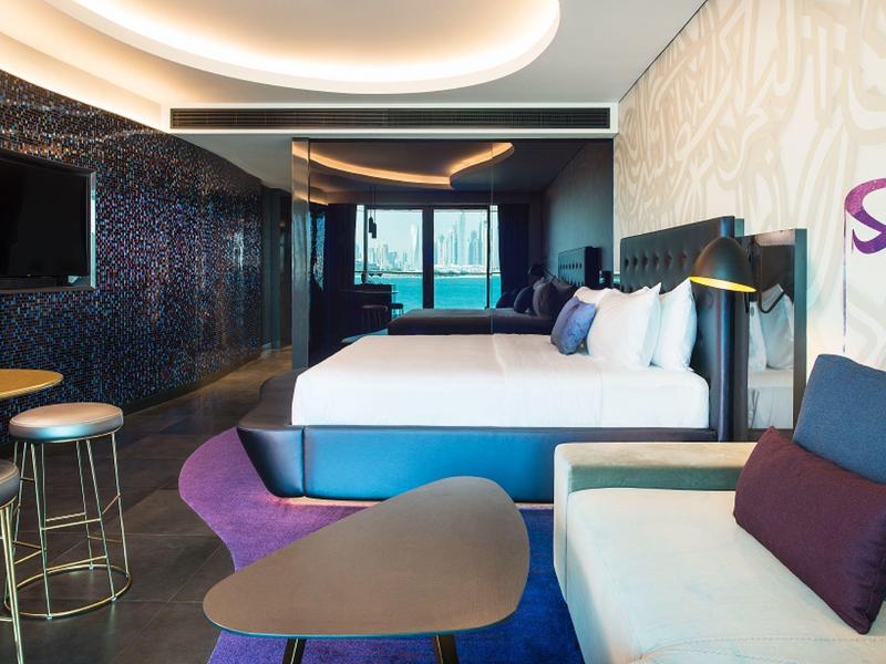 Modernes Hotelzimmer mit großem Bett, lila Akzenten, Sofa, Tisch und Blick auf Meer durch Fenster.