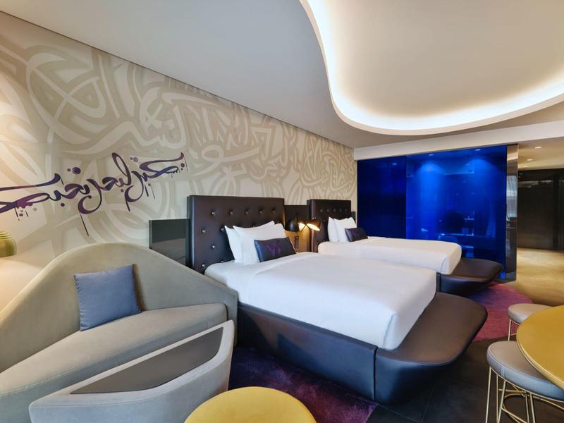 Modernes Hotelzimmer mit zwei Betten, stilvollen Möbeln, indirekter Deckenbeleuchtung und arabischer Wandkunst.