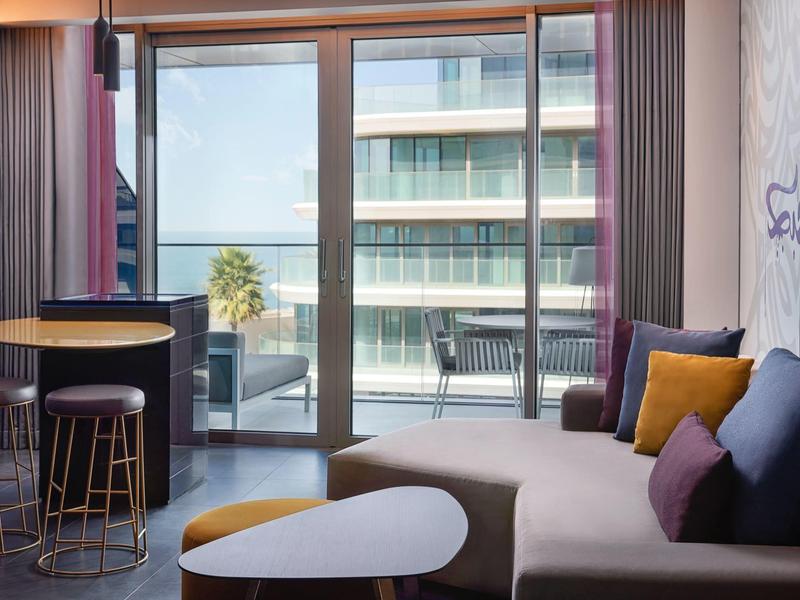 Moderner Wohnraum mit Sofa, bunten Kissen, Tisch und Balkon mit Blick auf moderne Gebäude.