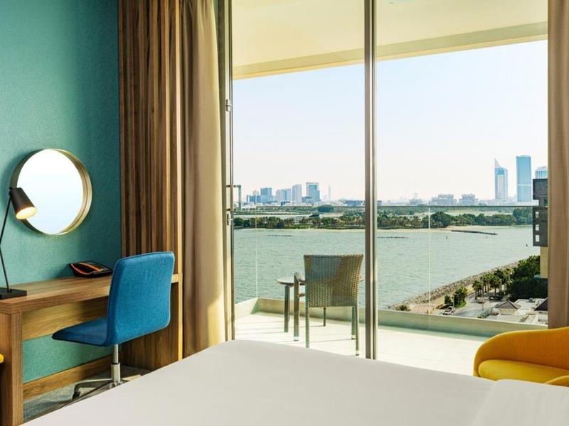 Modernes Hotelzimmer mit großen Fenstern, Blick auf Wasser, blauer Wand, Schreibtisch und farbigen Stühlen.