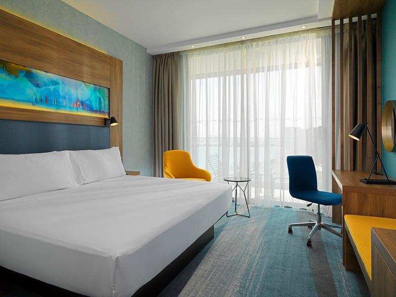 Modernes Hotelzimmer mit großem Bett, blauem Stuhl, gelbem Sessel und großem Fenster mit Vorhängen.