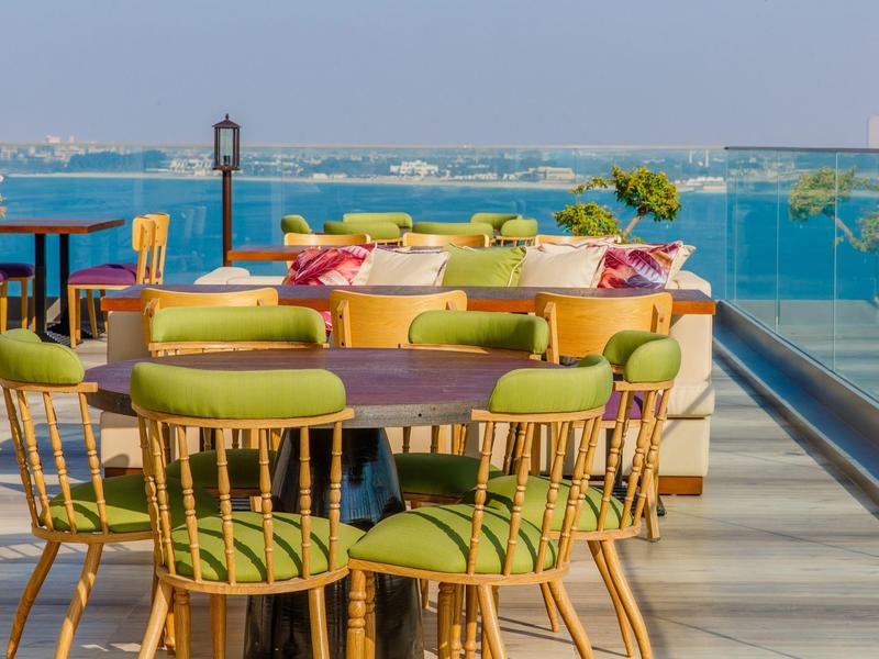Dachterrasse mit grünen Stühlen, Holztischen und Blick auf das blaue Meer bei klarem Himmel.