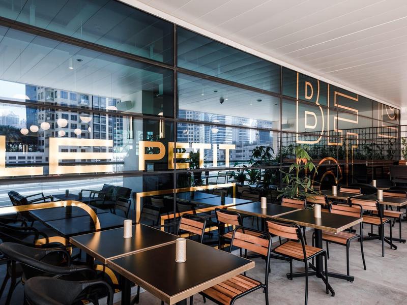 Café de rue moderne avec tables et chaises en bois devant une paroi vitrée de restaurant.