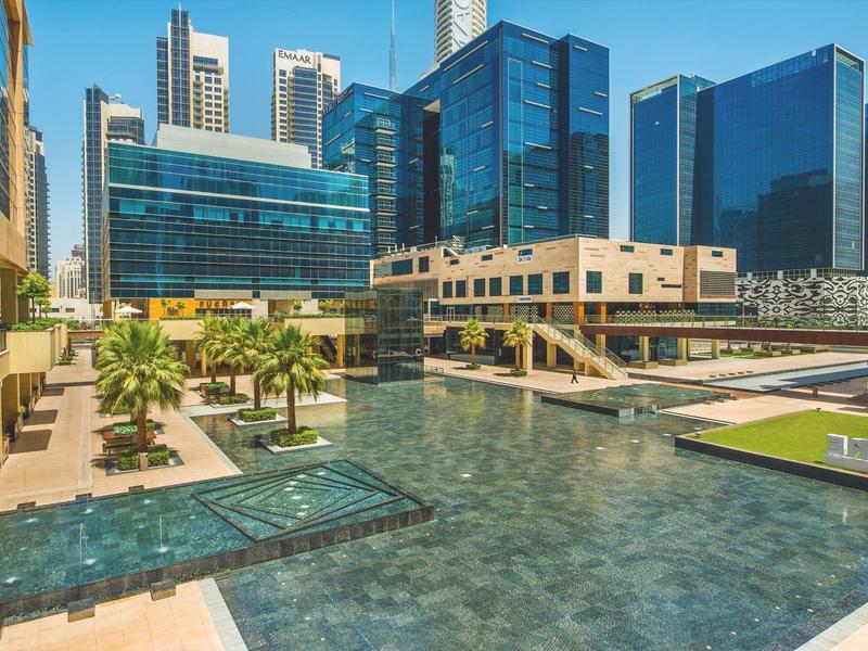 Complexe hôtelier moderne avec piscine et gratte-ciel en arrière-plan par une journée ensoleillée.