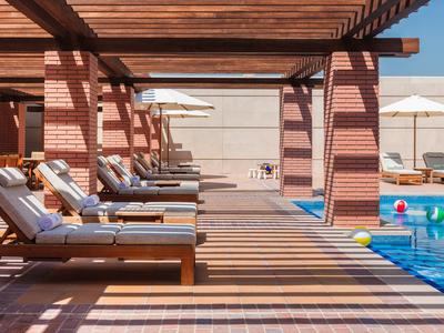Terraza soleada junto a la piscina con tumbonas bajo una pérgola de madera y pelotas de colores en el agua.