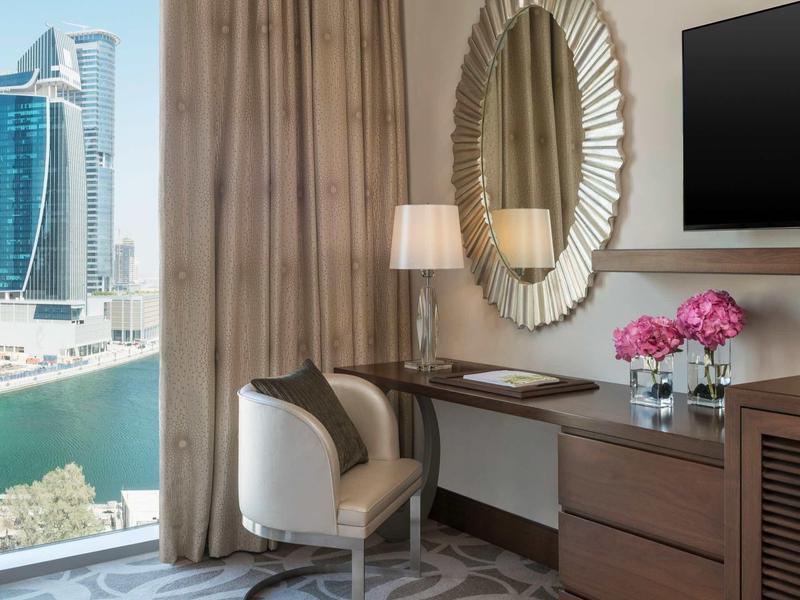 Lichte hotelkamer met bureau, stoel, ronde spiegel en uitzicht op water en wolkenkrabbers.