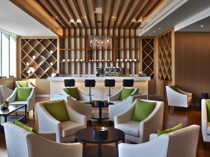 Lounge moderna di hotel con poltrone bianche, cuscini verdi, pannellature in legno e cantinetta.