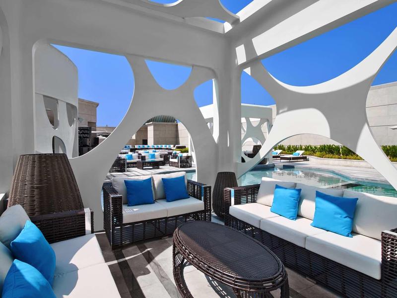 Area esterna moderna con divani bianchi, cuscini blu e lampade decorative sotto pergolato.