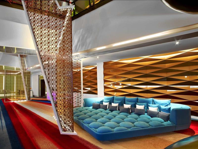 Area lounge moderna con divano imbottito blu, parete geometrica e tappeto rosso.