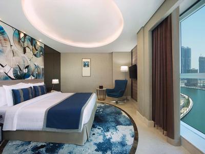 Habitación de hotel moderna con cama grande, silla azul y vista a un río y edificios.