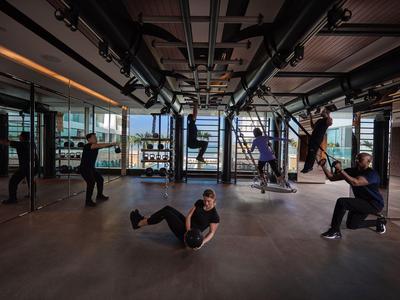 Diverse persone si allenano con bande TRX in una palestra moderna con pavimento in legno e soffitto a pannelli.