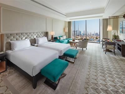 Prostorný hotelový pokoj se dvěma postelemi, zelenými podnožkami, pohodlnými křesly a velkými okny s výhledem na město.