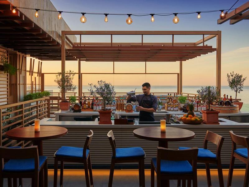 Večerní pohled na venkovní terasu s stoly, židlemi a rostlinami u moře.