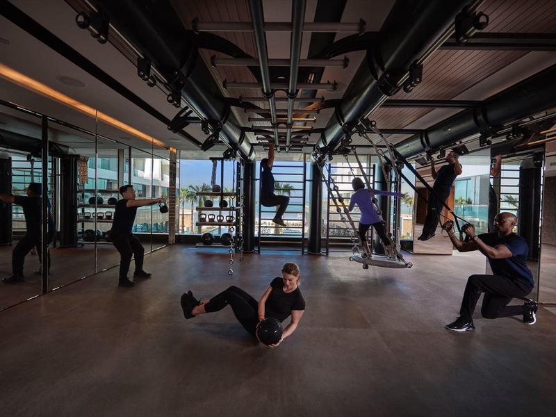 Několik lidí cvičí v moderní posilovně s velkými okny a výhledem ven.