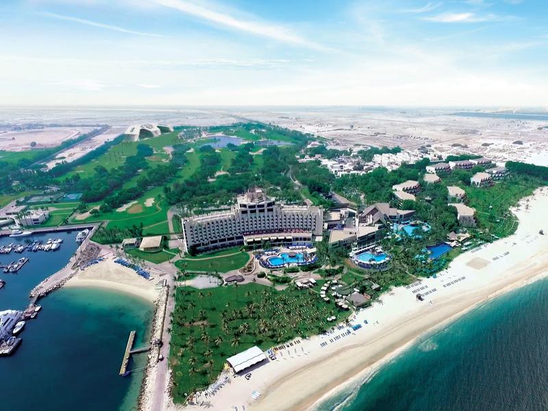 Luxury coastal hotel with pool, beach, and marina under a blue sky.