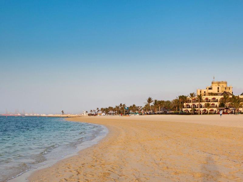 Wide sandy beach with calm sea and building at the edge under clear sky.