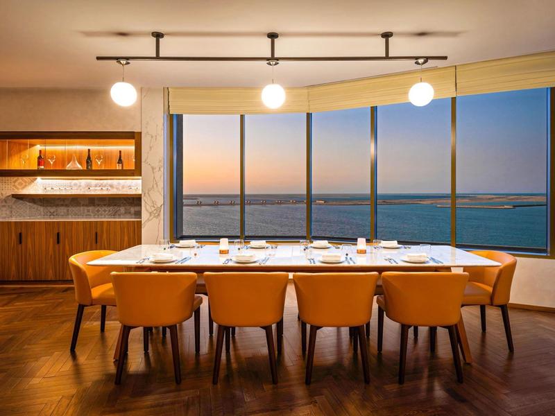 Modern dining room with large wooden table and orange chairs overlooking the sea at sunset.