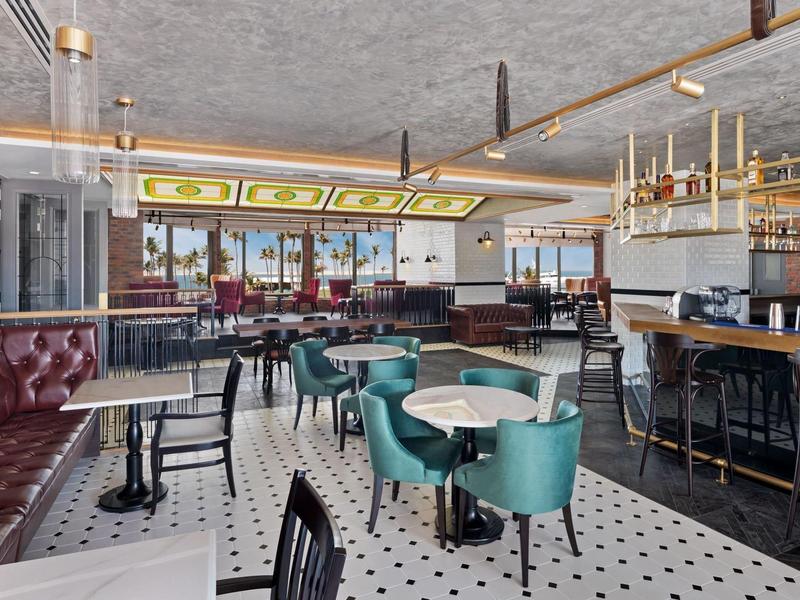 Modern hotel bar and lounge area with tables, chairs, and large windows overlooking the city.