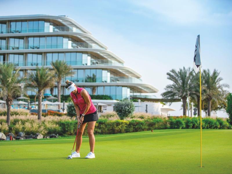 Golfer woman in red shirt playing on a manicured green near a modern hotel building.