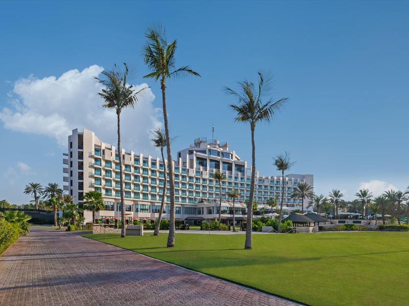 Modern hotel building with palm trees and a walkway in the foreground on a sunny day.