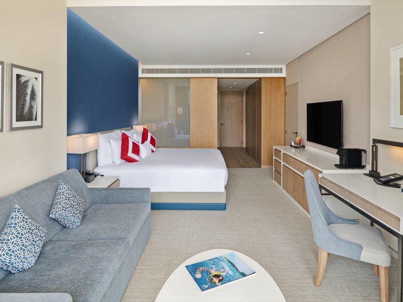 Modern furnished hotel room with bed, sofa, desk, and TV.