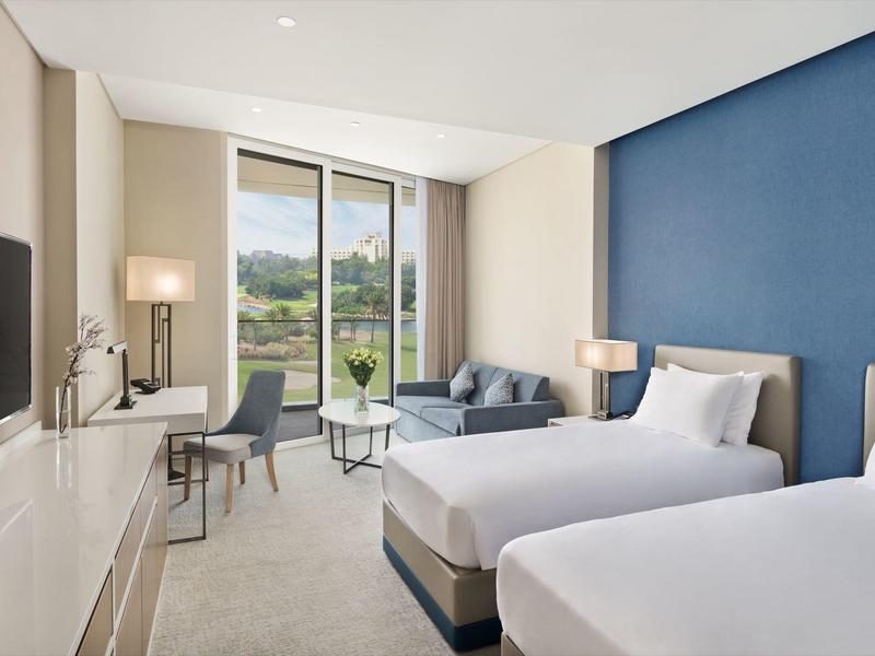 Modern hotel room with two single beds, desk, sofa, and large window offering a green view.