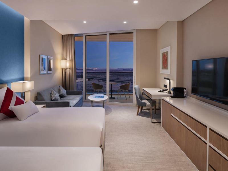 Modern hotel room with two beds, TV, desk, and city view at night.