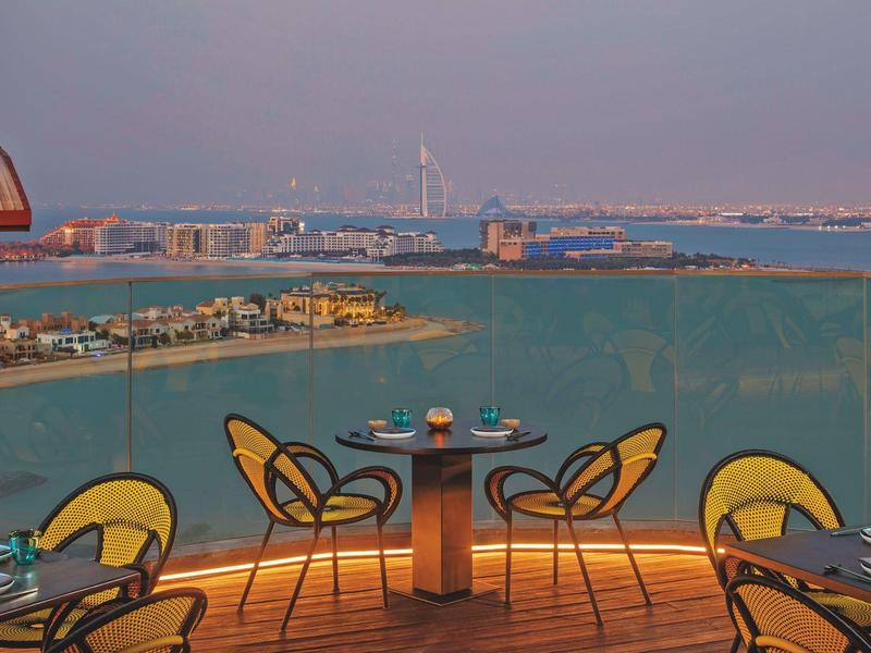 Balkon mit runden Tischen und gelb-schwarzen Stühlen, Blick auf Meer, Inseln und Stadt im Hintergrund.