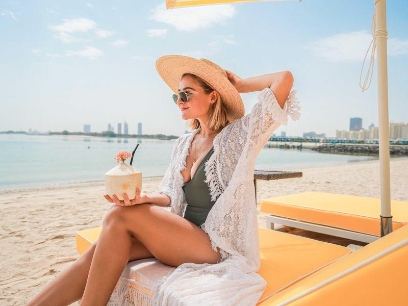 Frau im Strohhut und weißer Strandkleidung sitzt entspannt am sonnigen Sandstrand mit Cocktail.