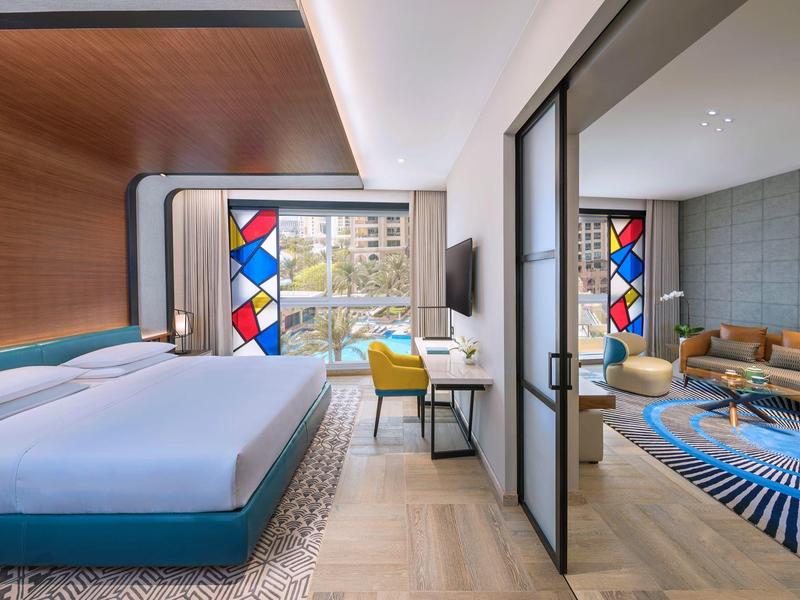 Modernes Hotelzimmer mit großem Bett, Schreibtisch, bunten Glasfenstern und angrenzendem Wohnzimmer.