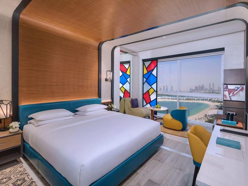 Modernes Hotelzimmer mit großem Bett, bunten Glasfenstern und Blick auf die Stadt und den Hafen.