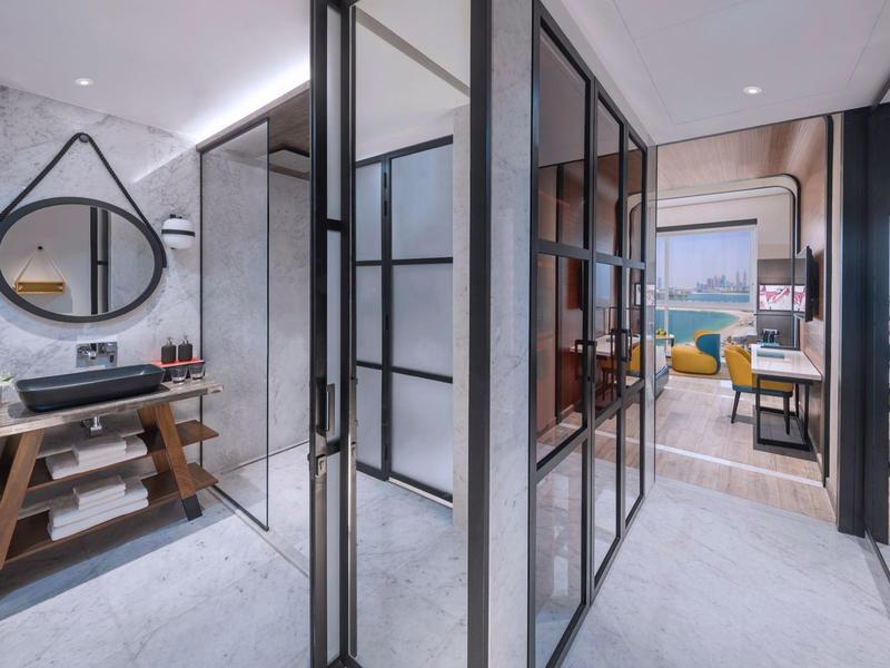 Moderner Flur mit Glaswänden, Bad mit Holzregal und Waschbecken, Blick ins Wohnzimmer mit gelbem Sofa.