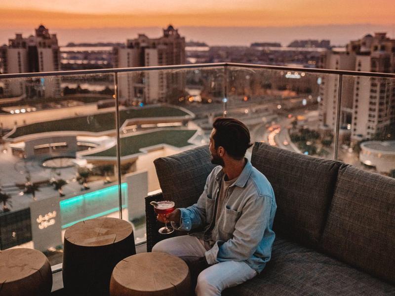 Mann sitzt abends auf Balkon mit Glasgeländer, Stadt und Sonnenuntergang im Hintergrund.