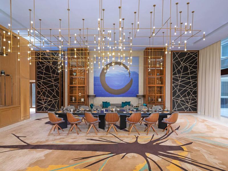 Moderner Tagungsraum mit langem Tisch, farbigen Stühlen, kunstvollem Teppich und hängenden Lichtern.