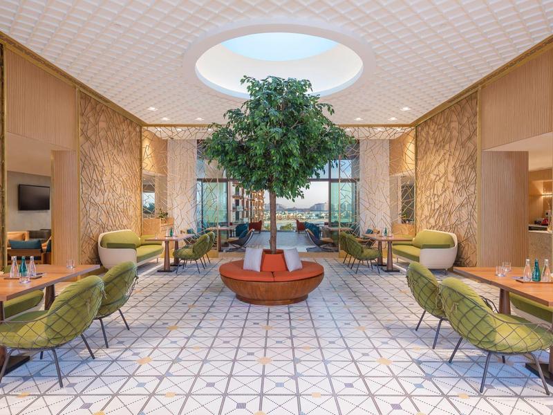 Großer, heller Wartebereich mit grünem Baum in der Mitte, grünen Sesseln und gemustertem Fliesenboden.